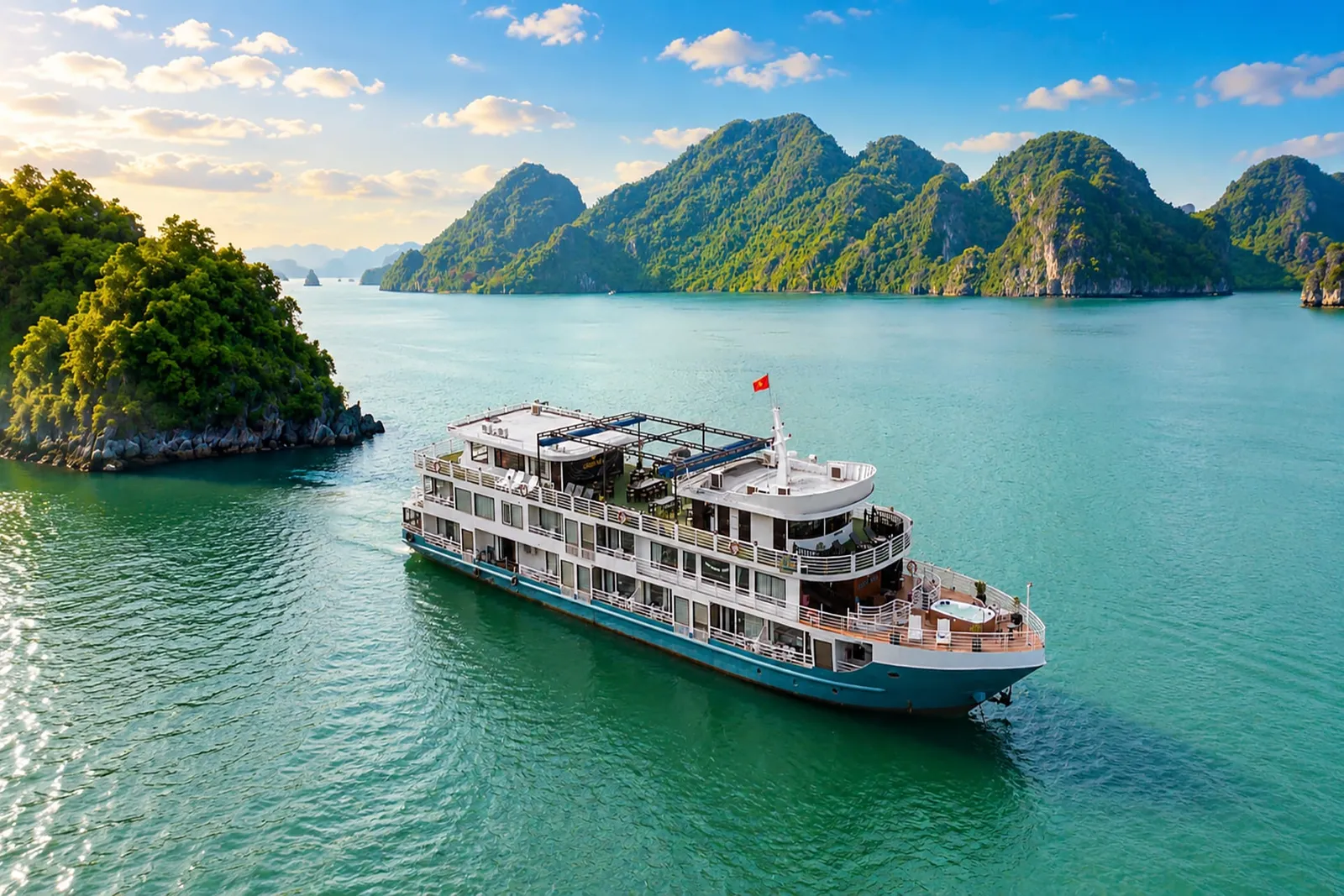 An aerial view of a multi-deck white and blue cruise boat flying a red flag, sailing on turquoise water past forested limestone karst islands under a blue sky with clouds.