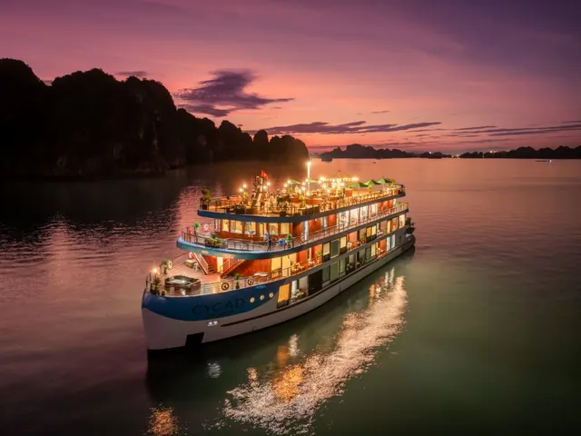 A multi-deck illuminated cruise ship with blue hull markings anchored in calm water at dusk with limestone karst formations silhouetted in the background.