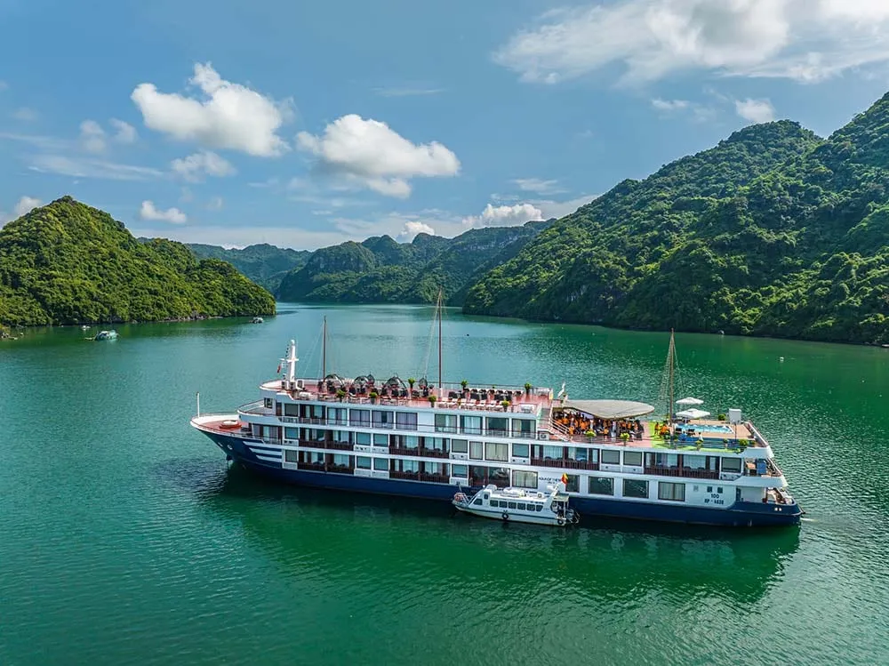A multi-deck cruise ship with blue and white painted hull sits in calm green water surrounded by steep forested limestone karst mountains.