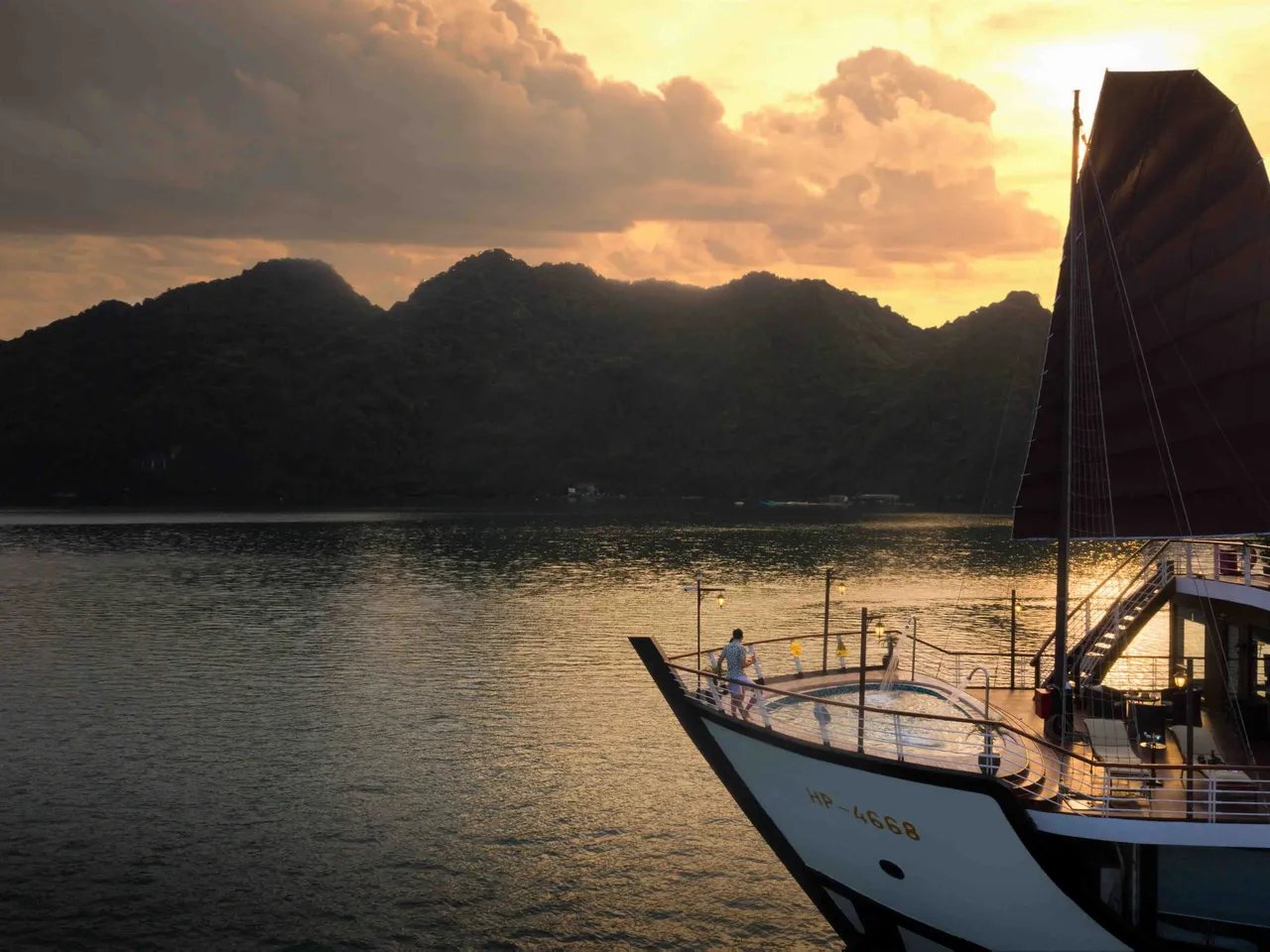 A cruise boat with dark sails and white hull bearing hull numbers anchored in calm water at sunset with limestone karst mountains in the background.