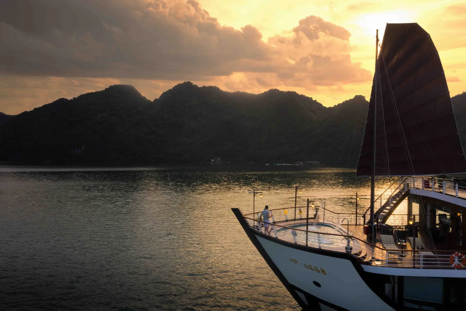 A cruise boat with dark sails and white hull bearing hull numbers anchored in calm water at sunset with limestone karst mountains in the background.