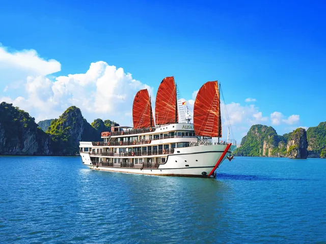 A white cruise ship with three red sails and the name 'ALISA' on its hull sailing through calm blue water surrounded by limestone karst islands.