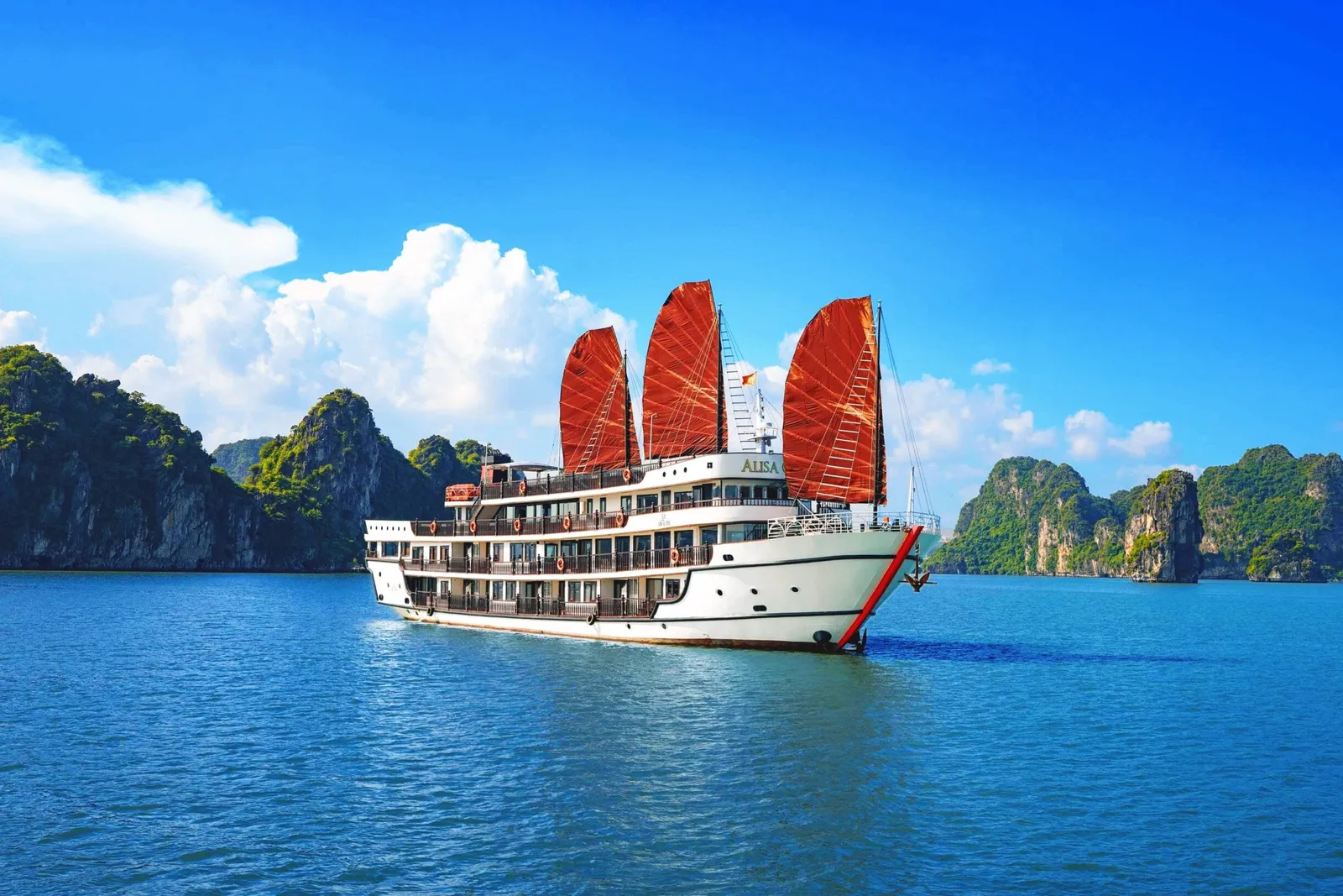 A white cruise ship with three red sails and the name 'ALISA' on its hull sailing through calm blue water surrounded by limestone karst islands.