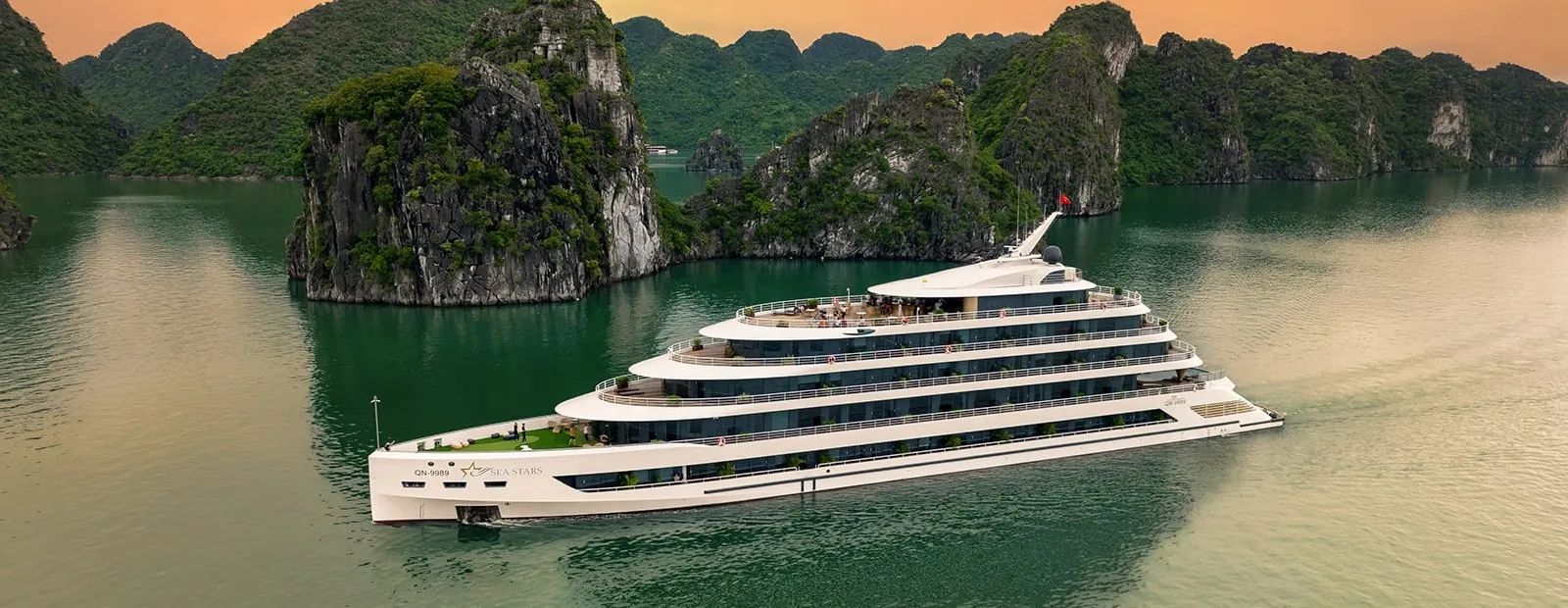 A large white multi-deck cruise ship with a star logo and name on its hull sails on green water past forested limestone karsts under an orange dusk sky, photographed from above and off to the side.
