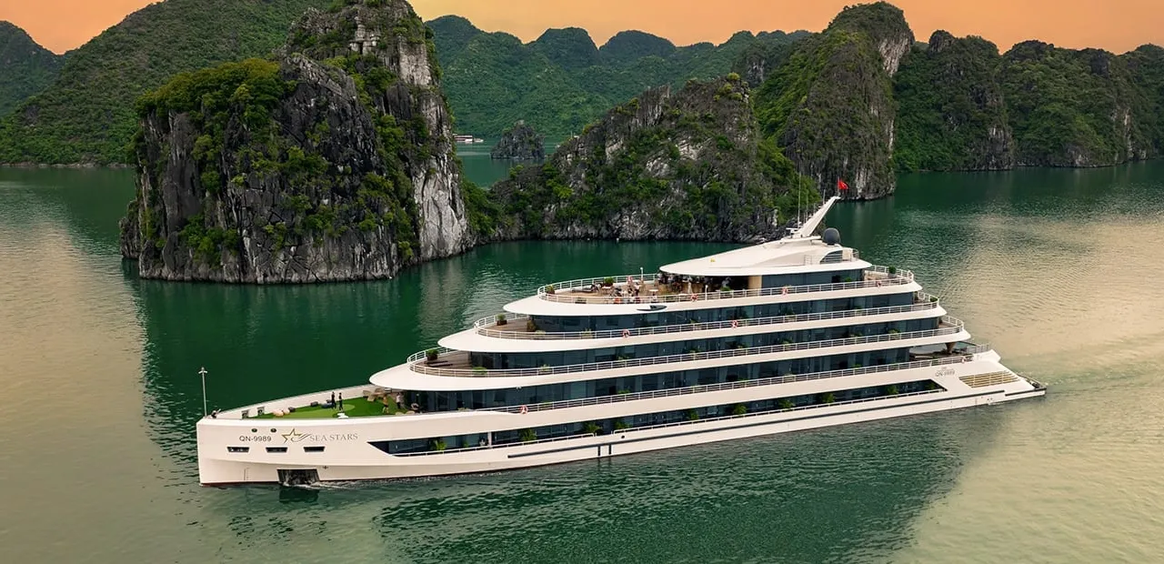 A large white multi-deck cruise ship with a star logo and name on its hull sails on green water past forested limestone karsts under an orange dusk sky, photographed from above and off to the side.