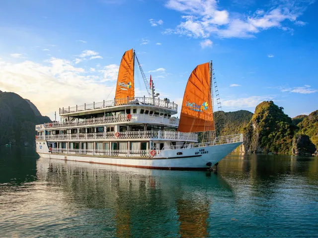A white multi-deck cruise ship with orange junk-style sails bearing a logo and hull registration text sits on calm green water in front of forested limestone karst islands under a blue sky.