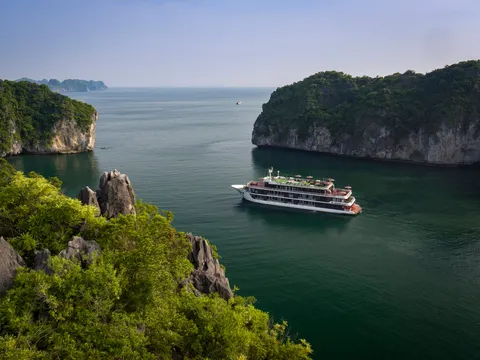 A multi-deck white cruise boat sits on calm green water between forested limestone islands, photographed from a high vantage point on a hillside.