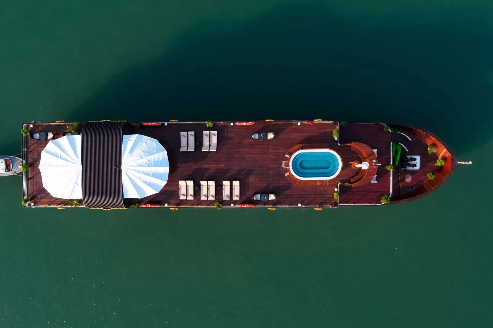 An aerial view of a wooden cruise boat with white sails, loungers, and a swimming pool on its deck floating in calm green water.