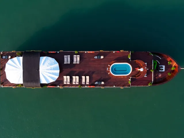 An aerial view of a wooden cruise boat with white sails, loungers, and a swimming pool on its deck floating in calm green water.