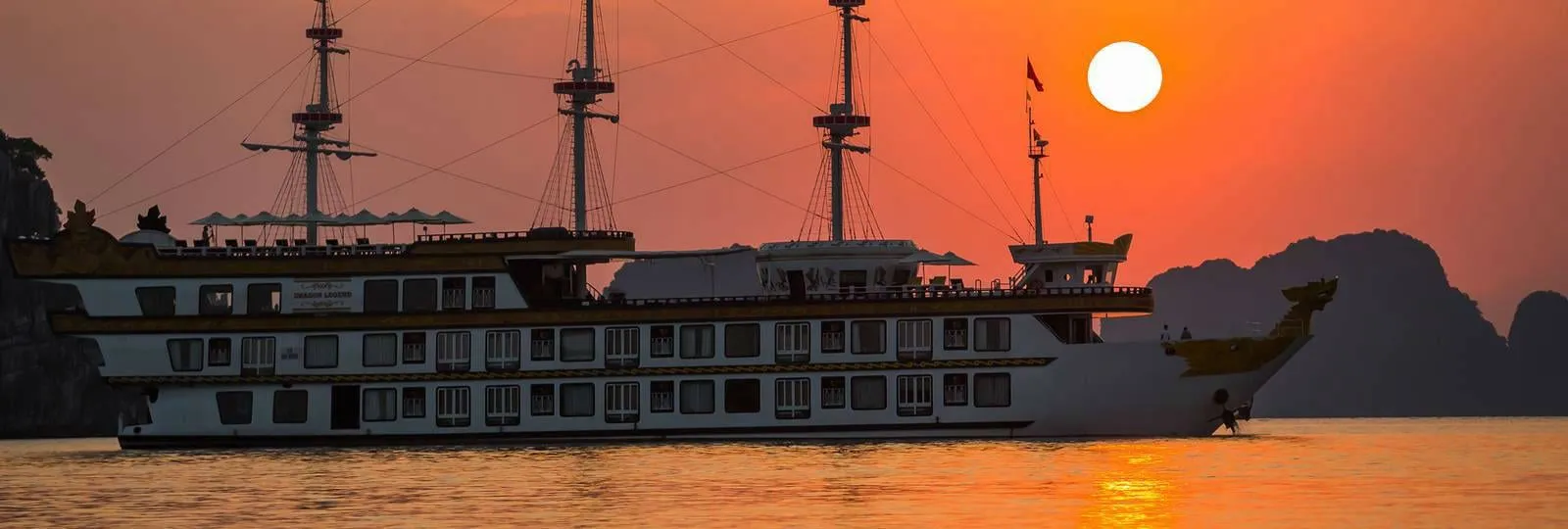 Dragon Legend side on against an orange sunset, three bare masts above the deck line and the sun sitting low behind
