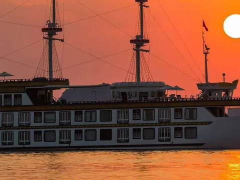 Dragon Legend side on against an orange sunset, three bare masts above the deck line and the sun sitting low behind