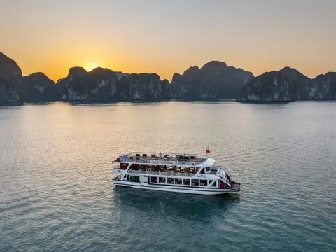 A white multi-deck cruise boat with a red flag sits on calm water at sunset with limestone karst formations in the background.