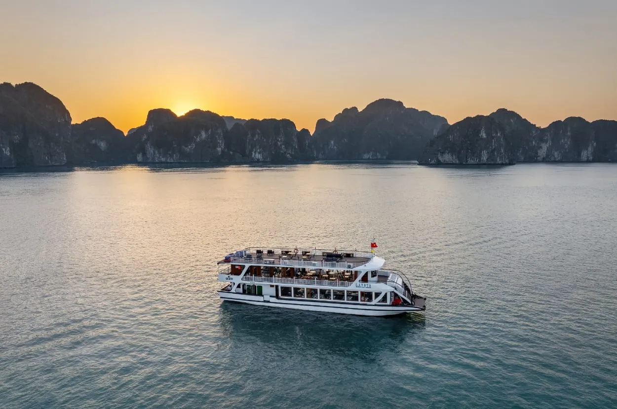 A white multi-deck cruise boat with a red flag sits on calm water at sunset with limestone karst formations in the background.