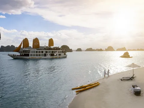A multi-deck cruise boat with orange junk sails, marked 'Emperor Cruises' on its hull, floats in a bay of limestone karsts at sunset, while two people in white stand on a sandy beach with yellow kayaks, loungers, an umbrella and a small table.