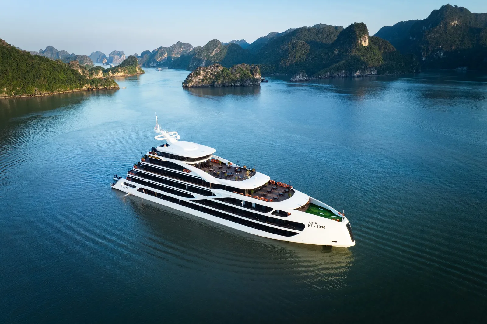 An aerial view of a white multi-deck cruise ship with dark horizontal bands sailing through calm blue water surrounded by limestone karst islands.