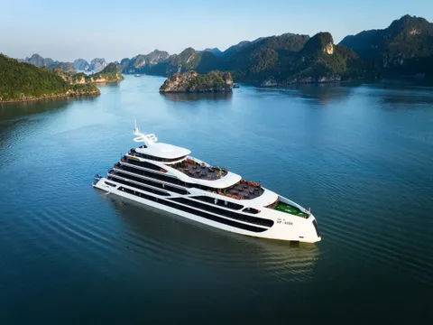 An aerial view of a white multi-deck cruise ship with dark horizontal bands sailing through calm blue water surrounded by limestone karst islands.