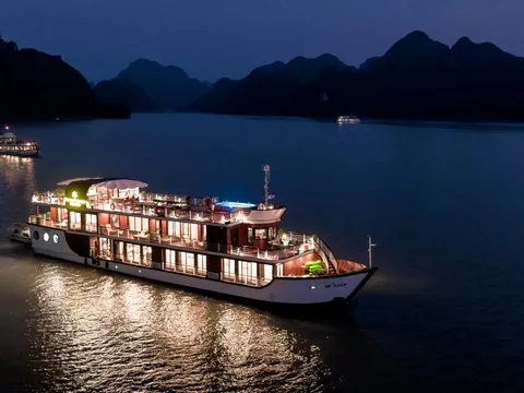 An illuminated cruise boat at twilight on calm water with dramatic karst limestone mountains silhouetted in the background.