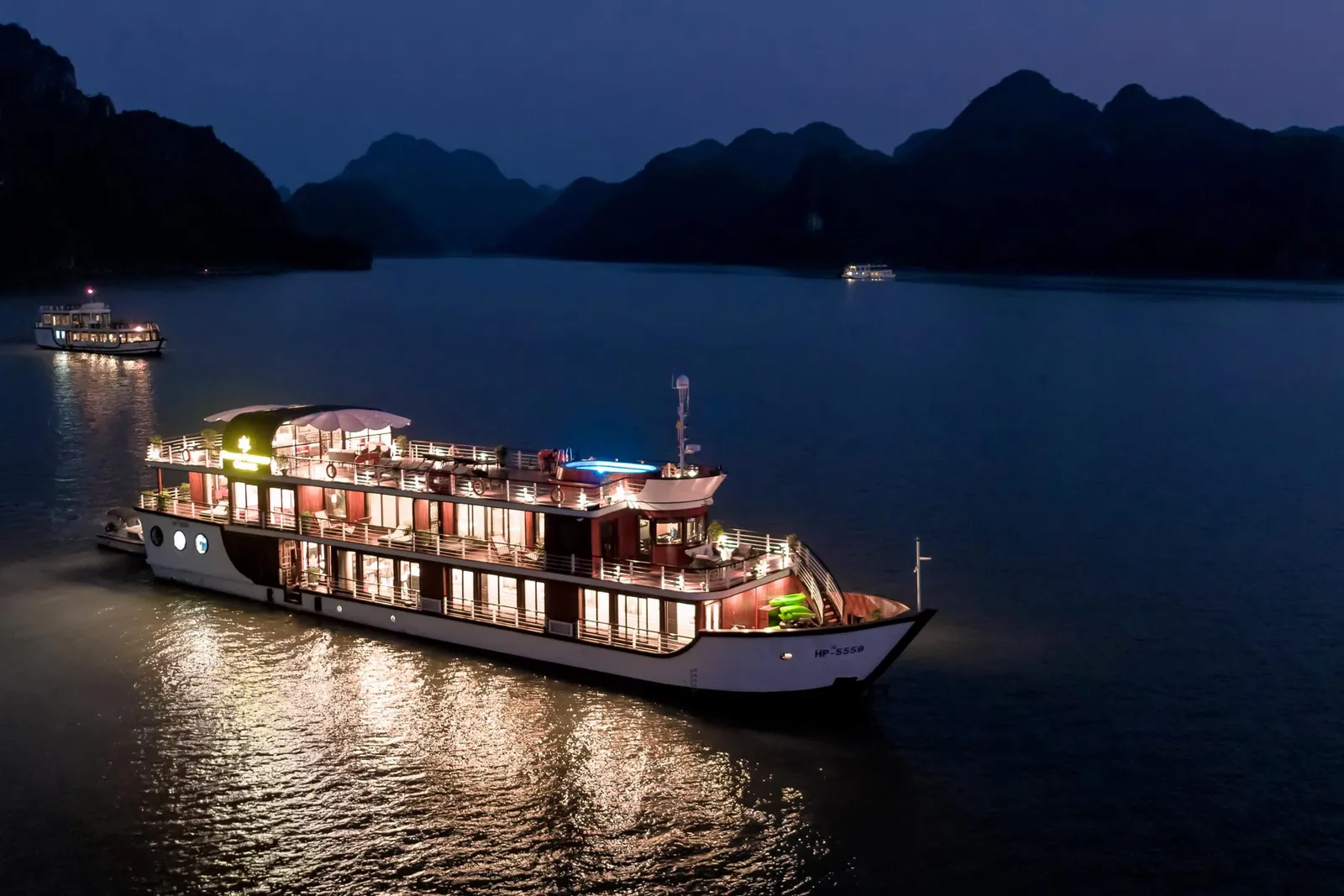 An illuminated cruise boat at twilight on calm water with dramatic karst limestone mountains silhouetted in the background.