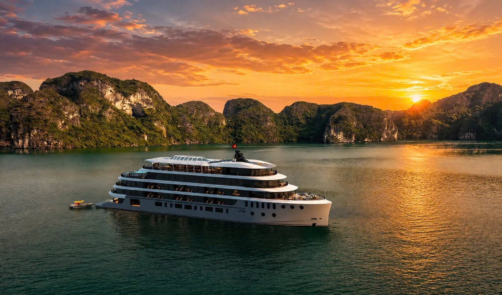 A multi-deck white cruise ship with a small red flag sits on calm green water in front of forested limestone karsts at sunset, with a small tender boat nearby.
