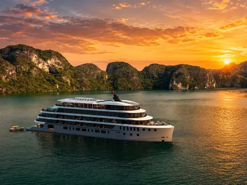 A multi-deck white cruise ship with a small red flag sits on calm green water in front of forested limestone karsts at sunset, with a small tender boat nearby.