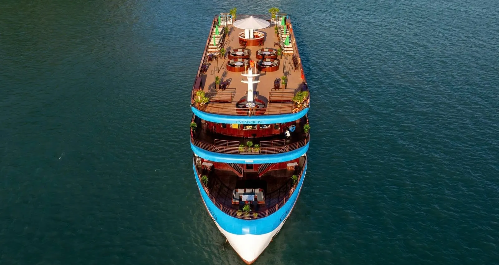 Aerial top-down view of a blue and white multi-deck cruise ship with wooden decks, round seating booths and an umbrella on the top deck, sailing on dark green water.