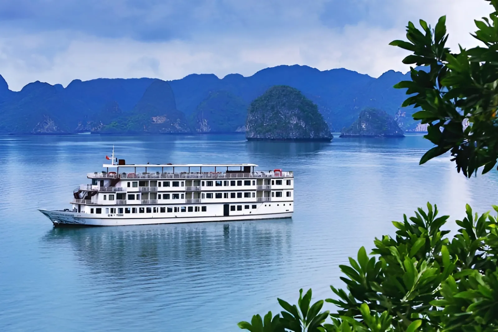 A white multi-deck cruise ship sails in calm blue water with dramatic limestone karst islands in the background, framed by green foliage in the foreground.