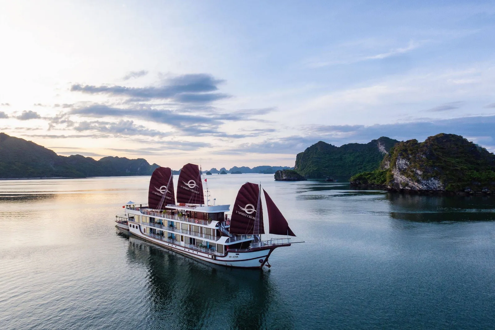 An aerial view of a white multi-deck cruise ship with three dark maroon sails bearing a logo and 'V'Spirit Cruises' text, floating on calm water near green limestone islands at sunset.