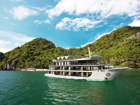 A white multi-deck cruise ship with text on its upper deck sails in green water in front of forested limestone karst islands under blue sky.