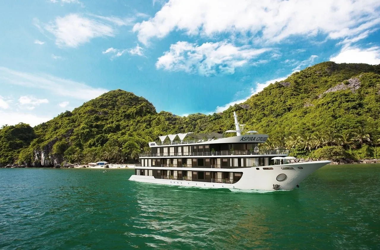 A white multi-deck cruise ship with text on its upper deck sails in green water in front of forested limestone karst islands under blue sky.