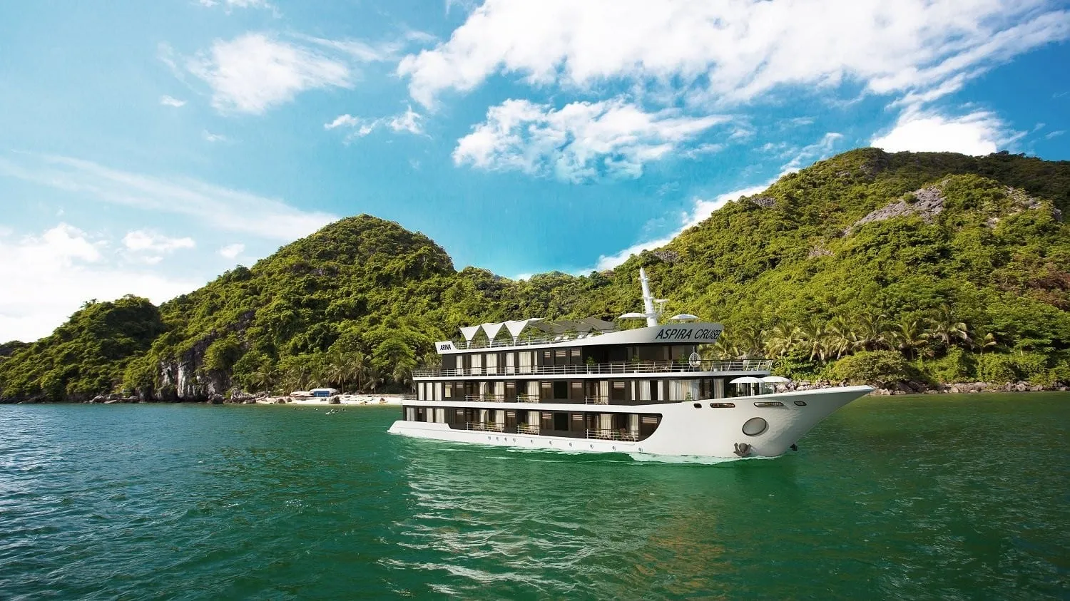 A white multi-deck cruise ship with text on its upper deck sails in green water in front of forested limestone karst islands under blue sky.