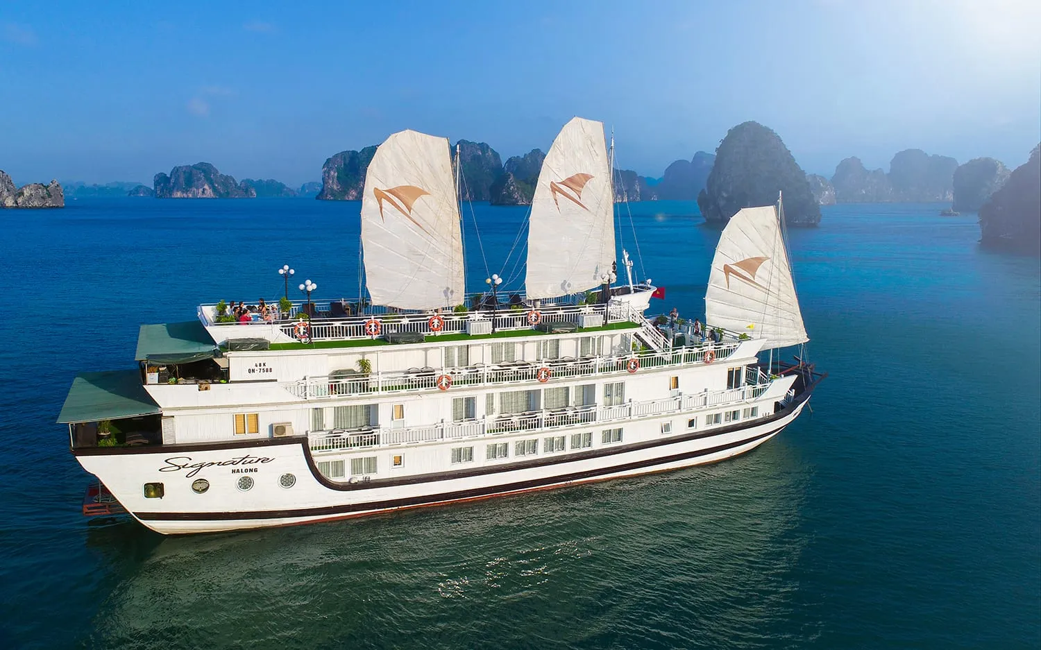A large white multi-deck cruise junk with three white sails bearing a bird logo, named 'Signature Halong' painted on the hull, sailing among karst limestone islands.