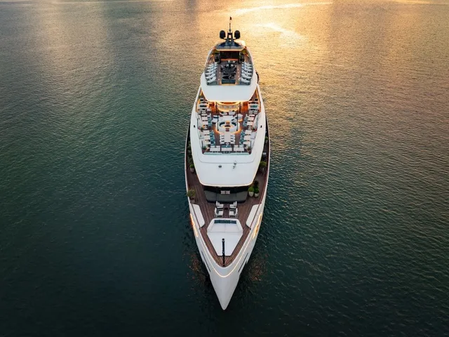 Aerial top-down view of a large white multi-deck cruise yacht with teak decks, seating and potted plants, on calm water lit by golden sunset light, with no land visible.