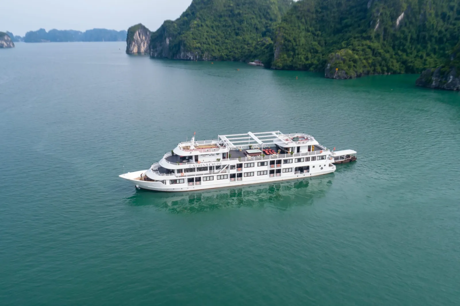 An aerial view of a white multi-deck cruise ship anchored in calm turquoise water surrounded by dramatic limestone karst islands covered in dense green vegetation.