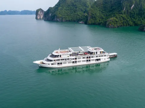 An aerial view of a white multi-deck cruise ship anchored in calm turquoise water surrounded by dramatic limestone karst islands covered in dense green vegetation.