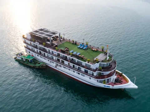 An aerial view of a large multi-deck white cruise ship with a green artificial-turf sun deck and blue loungers, with a small green service boat alongside it on calm green-teal water, with sun glare at upper left.