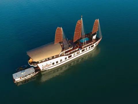 An aerial drone view of a white three-masted junk-style cruise boat with brown sails and the name 'Red Dragon' painted on its hull, with a smaller tender boat and a speedboat alongside on calm dark teal water.