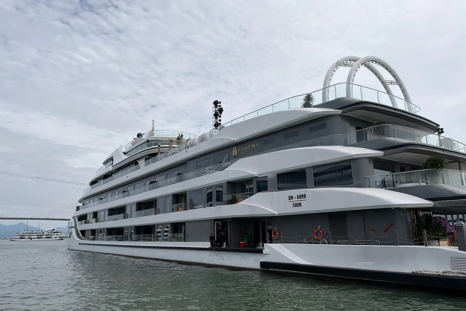 A large multi-deck white and grey cruise ship with the name 'Athena' on its hull and registration 'QN-6899' moored on grey-green water, with a bridge and another vessel in the distance under an overcast sky.