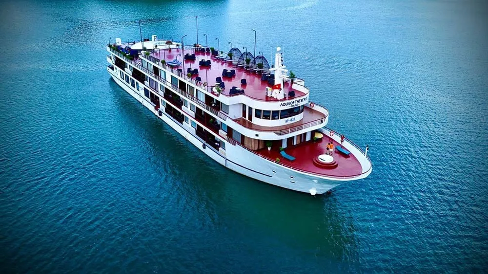 An aerial view of a white multi-deck cruise ship with pink or red upper decks floating in calm turquoise water.
