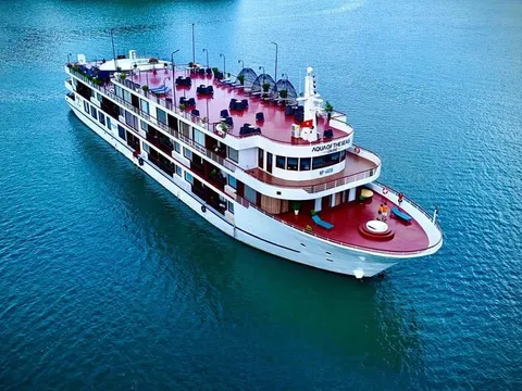 An aerial view of a white multi-deck cruise ship with pink or red upper decks floating in calm turquoise water.