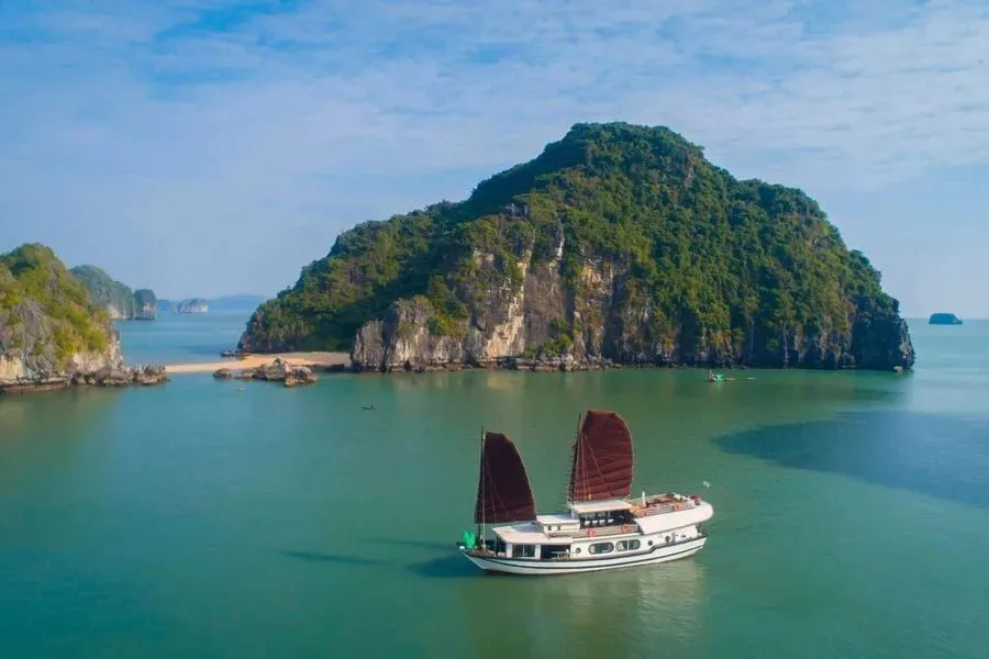 An aerial view of a white two-deck junk boat with dark red sails on calm green water in front of a forested limestone island with a small sandy beach.