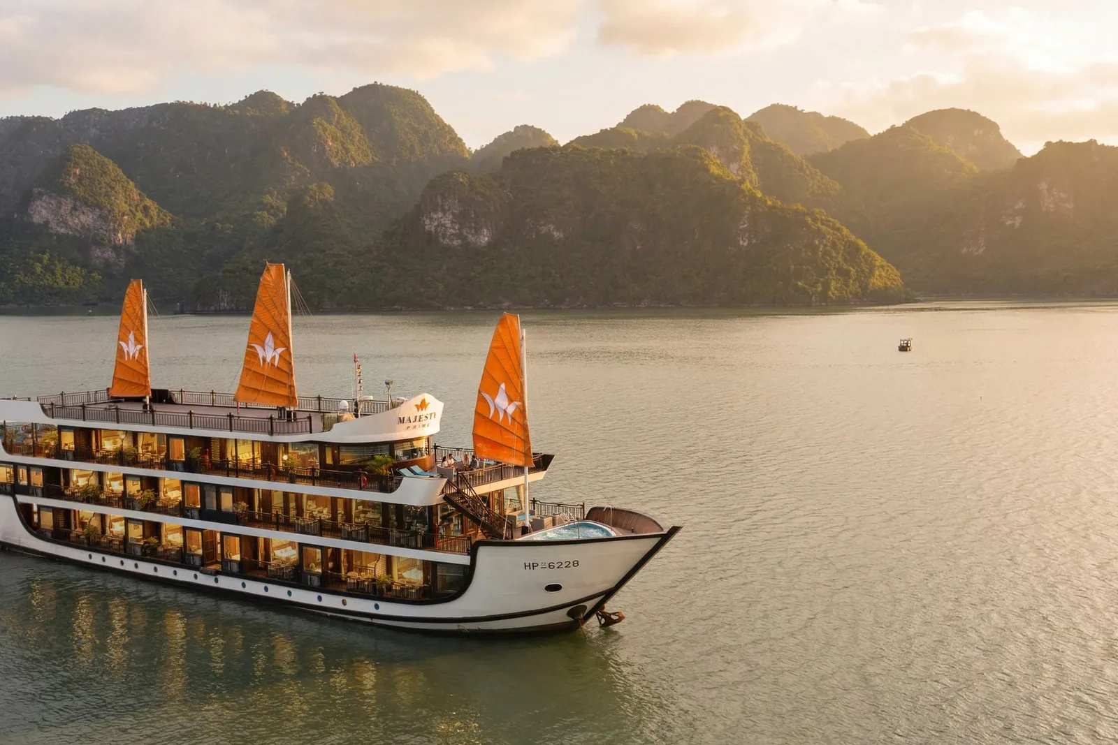 A white cruise boat with three orange sails and illuminated cabins floats on calm water with dramatic limestone karst mountains in the background at sunset.