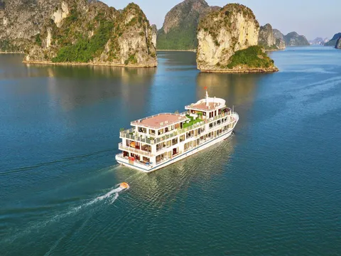 An aerial view of a multi-deck white cruise boat with a small orange tender in its wake, sailing on calm green-blue water past limestone karst islands.
