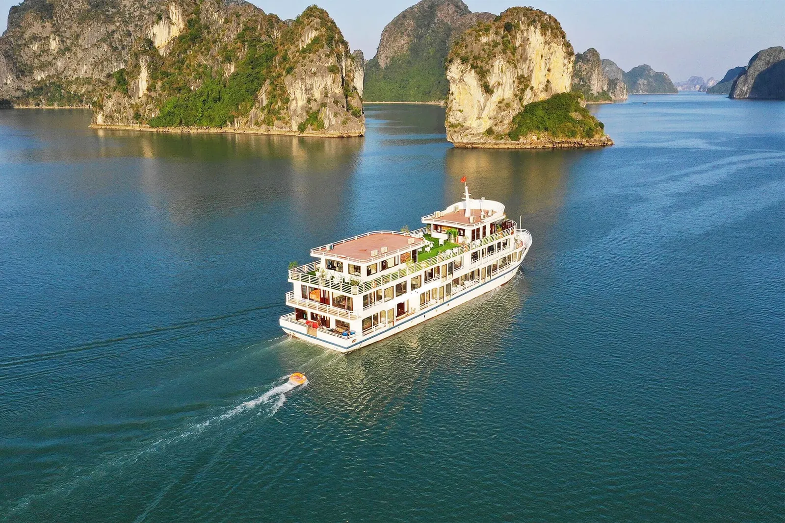 An aerial view of a multi-deck white cruise boat with a small orange tender in its wake, sailing on calm green-blue water past limestone karst islands.