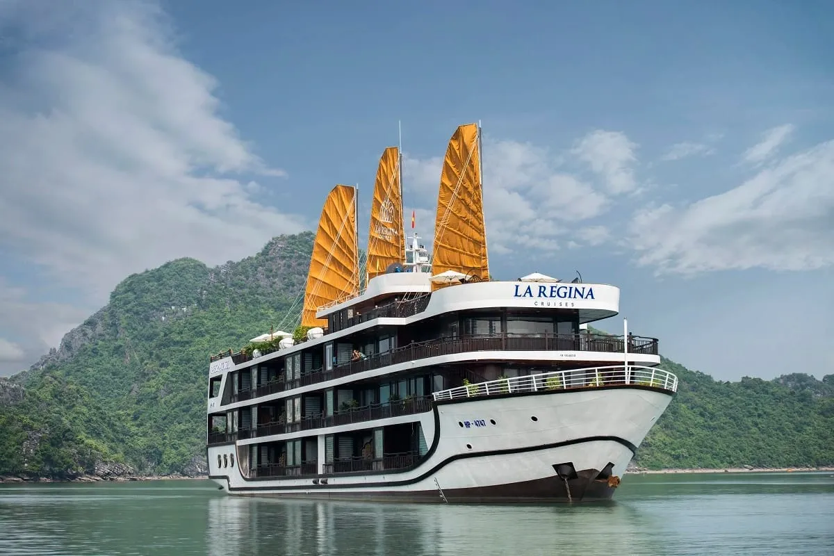 A multi-deck white cruise ship with three orange sails and 'La Regina Cruises' lettering on its upper deck sits on calm green water in front of forested limestone karst hills under a blue sky with clouds.