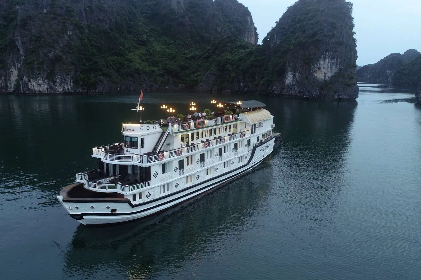 A white multi-deck cruise ship with lights on sails through calm water surrounded by tall limestone karst formations at dusk.
