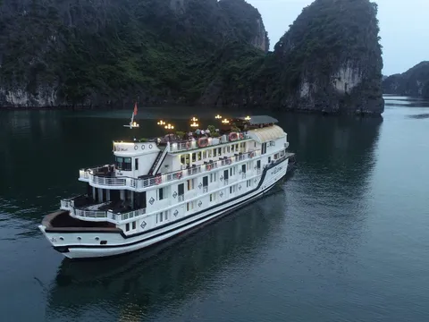 A white multi-deck cruise ship with lights on sails through calm water surrounded by tall limestone karst formations at dusk.
