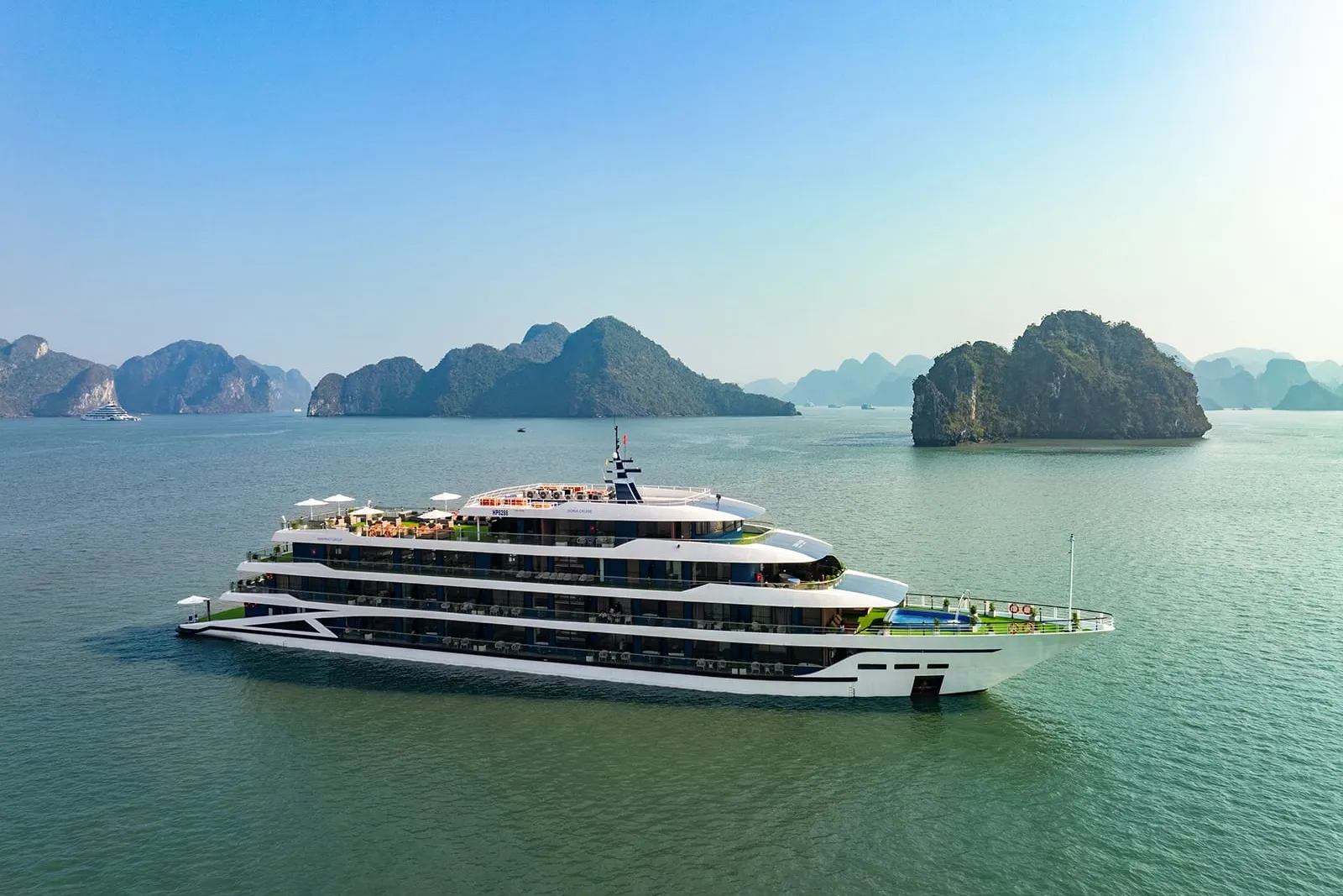A large modern white cruise ship with multiple decks floats in calm green water surrounded by dramatic limestone karst islands.