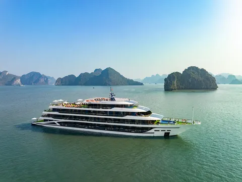 A large modern white cruise ship with multiple decks floats in calm green water surrounded by dramatic limestone karst islands.