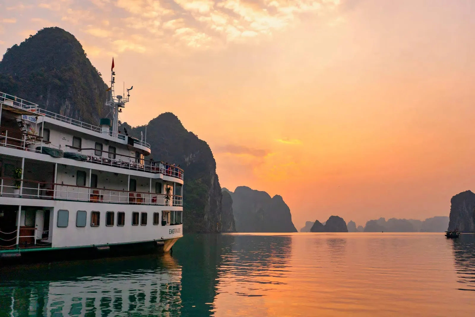 A white multi-deck cruise boat with the name 'Emeraude' on its hull sits on calm water beside limestone karst islands under an orange sunset sky, with a small boat in the distance.