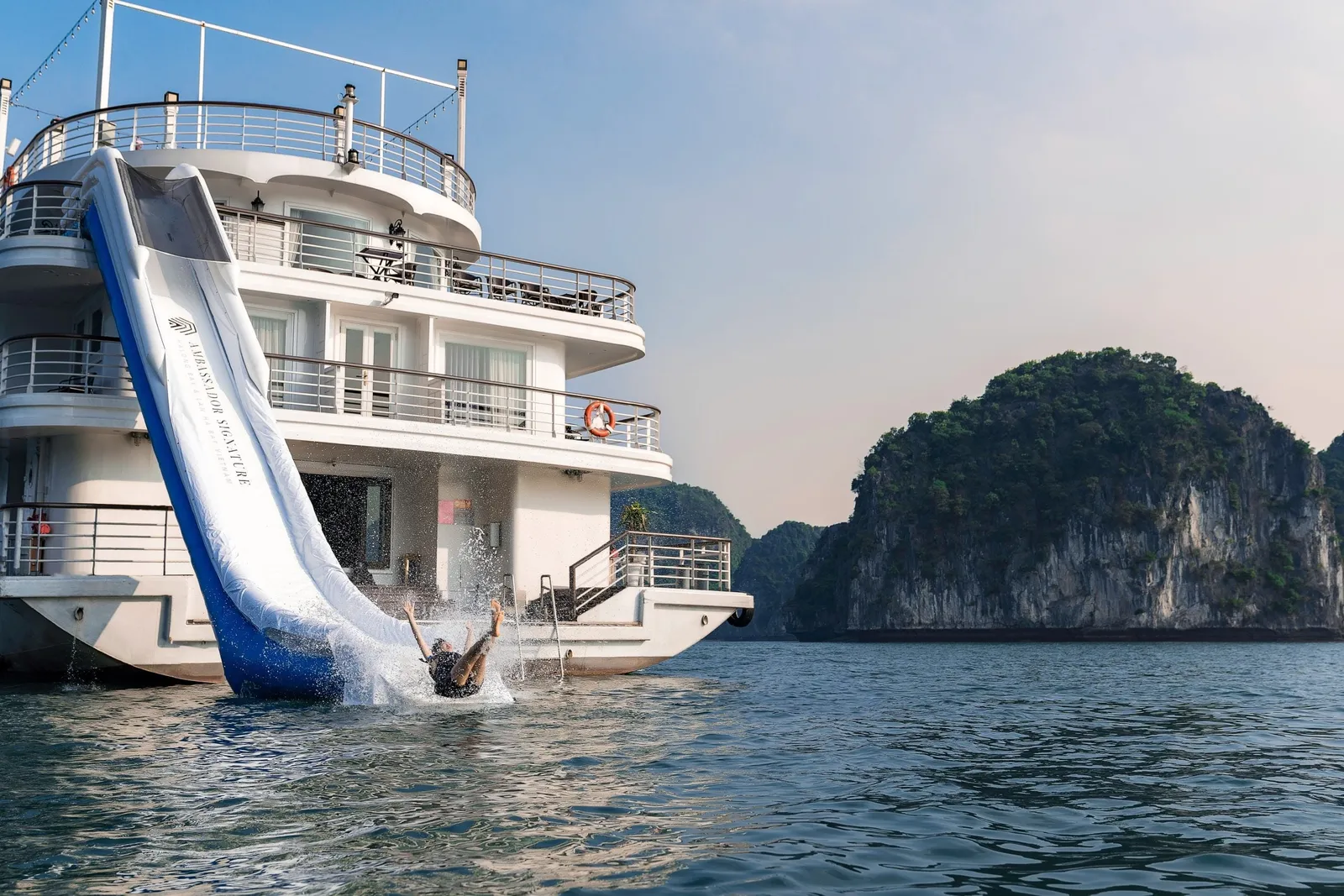 The stern of a white multi-deck cruise ship with an inflatable water slide printed 'Ambassador Signature', a person splashing into the water, and a limestone karst island beside it.